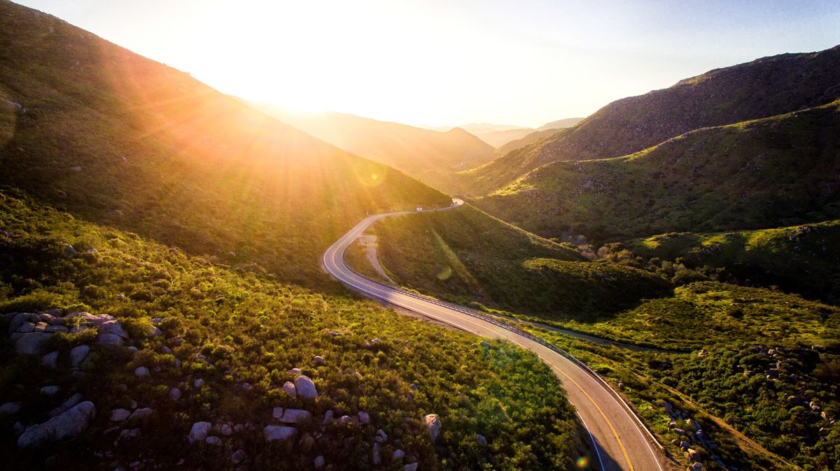 A landscape photo depicting a road between two lush hills and a sun set in the background