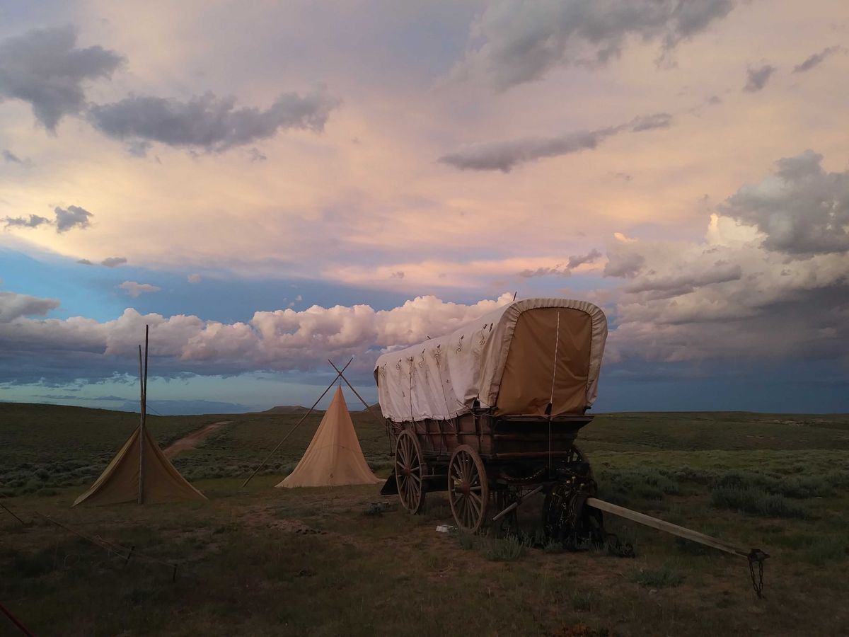 A photo of a covered wagon on a grassy plane with tents nearby